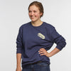 A woman stands smiling with one hand on her hip, wearing the New Duds Butter Long Sleeve T-shirt in navy blue heavyweight cotton, featuring the BUTTER logo on the chest, against a plain white background.