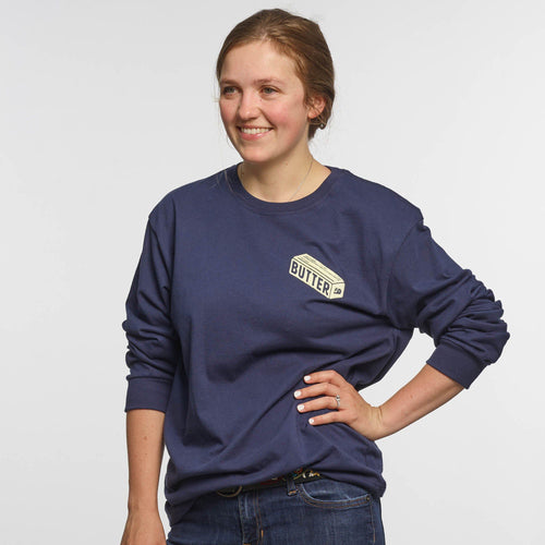 A woman stands smiling with one hand on her hip, wearing the New Duds Butter Long Sleeve T-shirt in navy blue heavyweight cotton, featuring the BUTTER logo on the chest, against a plain white background.