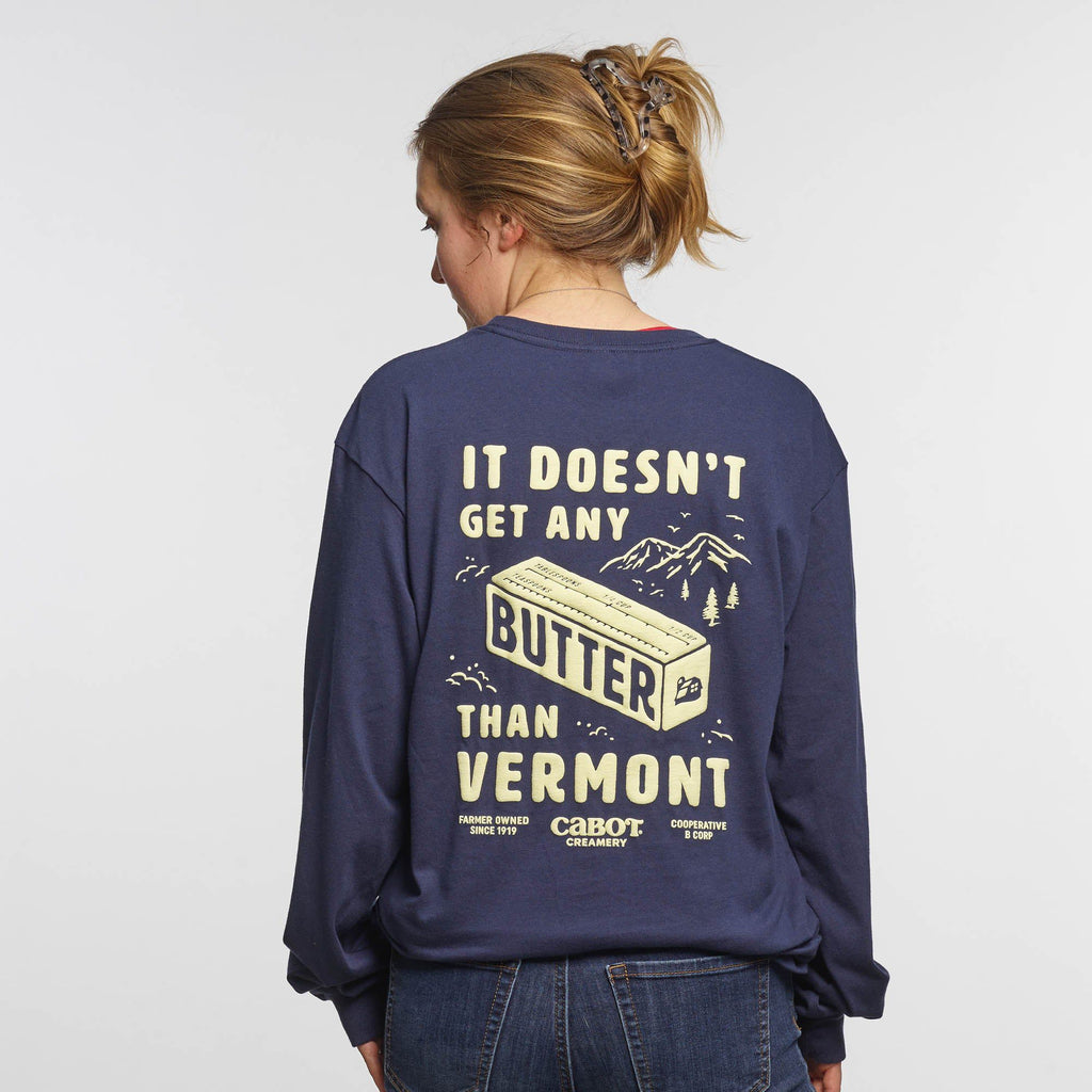 A person with light brown hair in a ponytail wears the New Duds Butter Long Sleeve T-shirt—a navy heavyweight cotton shirt featuring yellow It doesn’t get any butter than Vermont text and a butter illustration.