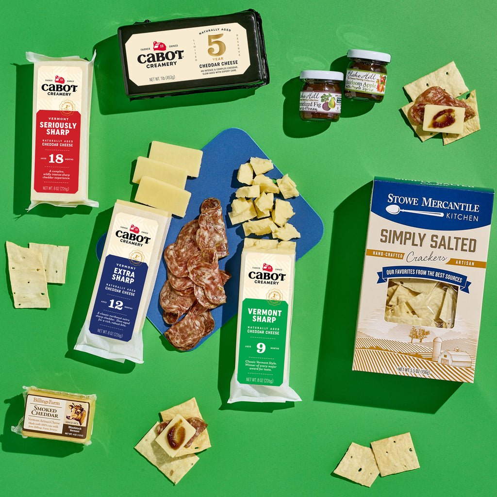 The Classic Vermont Charcuterie Gift Box by Cabot Creamery, displayed against a green backdrop, includes Vermont cheddar cheese, other Cabot cheeses, salami, Simply Salted crackers, chocolate squares, and small jars of jam.