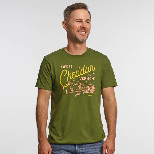 A man with short brown hair and a beard smiles in the New Duds Life is Cheddar in Vermont T-shirt, made of ring-spun cotton and featuring yellow text over a rustic farm scene with mountains, trees, cows, and a barn.