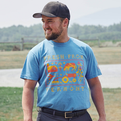 A man in a black cap and a New Duds Cabot x Always With Honor T-shirt with colorful Fresh From Vermont graphics stands smiling outdoors, grassy area beneath him and blurred trees and mountains in the background.