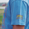A person wears a New Duds Cabot x Always With Honor T-shirt in light blue with colorful front patterns and a stitched Cabot Award Winner patch on the sleeve. The outdoor background is blurred.