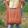 A person in a yellow shirt and blue jeans stands outdoors holding the rust-colored Cabot x Always With Honor Baggu reusable shopping bag by Baggu, with green plants and grass visible in the background.