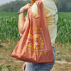 A person in a yellow shirt and smartwatch stands in a grassy field, carrying the Cabot x Always With Honor Baggu pink reusable bag by Baggu, featuring yellow pixel designs and FARM VERMONT text.