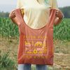 Someone in a yellow shirt holds the Cabot x Always With Honor Baggu by Baggu, a brown reusable bag featuring yellow cross-stitch designs and Fresh From Vermont text. They stand outdoors in a field.