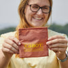 A smiling person with glasses holds up the Cabot x Always With Honor Baggu, a small reddish pouch by Baggu with CABOT ALWAYS WITH HONOR in yellow text. The outdoor background is slightly blurred.