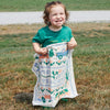 A smiling child with curly hair stands on grass wearing a green shirt, holding up the Cabot x Always With Honor Bandanna by New Duds, featuring “FRESH FROM...” text and colorful designs. A gravel path is visible in the background.
