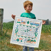 A young boy stands on grass in a rural setting holding the Cabot x Always With Honor Bandanna by New Duds, featuring colorful farm illustrations, trucks, cows, and the words Fresh from Vermont and Since 1919 Cabot.