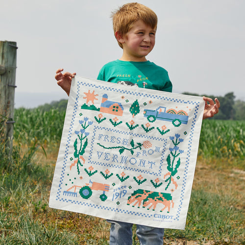 A young boy stands on grass in a rural setting holding the Cabot x Always With Honor Bandanna by New Duds, featuring colorful farm illustrations, trucks, cows, and the words Fresh from Vermont and Since 1919 Cabot.