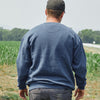 A man wearing the New Duds Cabot x Always With Honor Crewneck and a black cap stands with his back to the camera in a field of green crops, trees in the background.