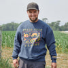 A smiling man in a blue Cabot x Always With Honor Crewneck by New Duds stands in front of a green field on a cloudy day.