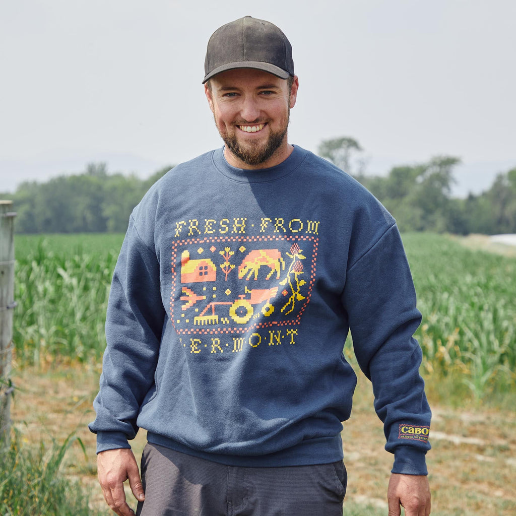 A smiling man in a blue Cabot x Always With Honor Crewneck by New Duds stands in front of a green field on a cloudy day.