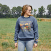 A woman stands in a green field under a hazy sky, wearing glasses, jeans, and the Cabot x Always With Honor Crewneck by New Duds with colorful farm-themed graphics. Trees line the background.