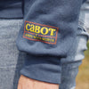 A close-up of the New Duds Cabot x Always With Honor Crewneck shows a blue sleeve with a stitched CABOT x ALWAYS WITH HONOR patch in yellow, red, and green. A hand and part of light blue jeans are also visible.