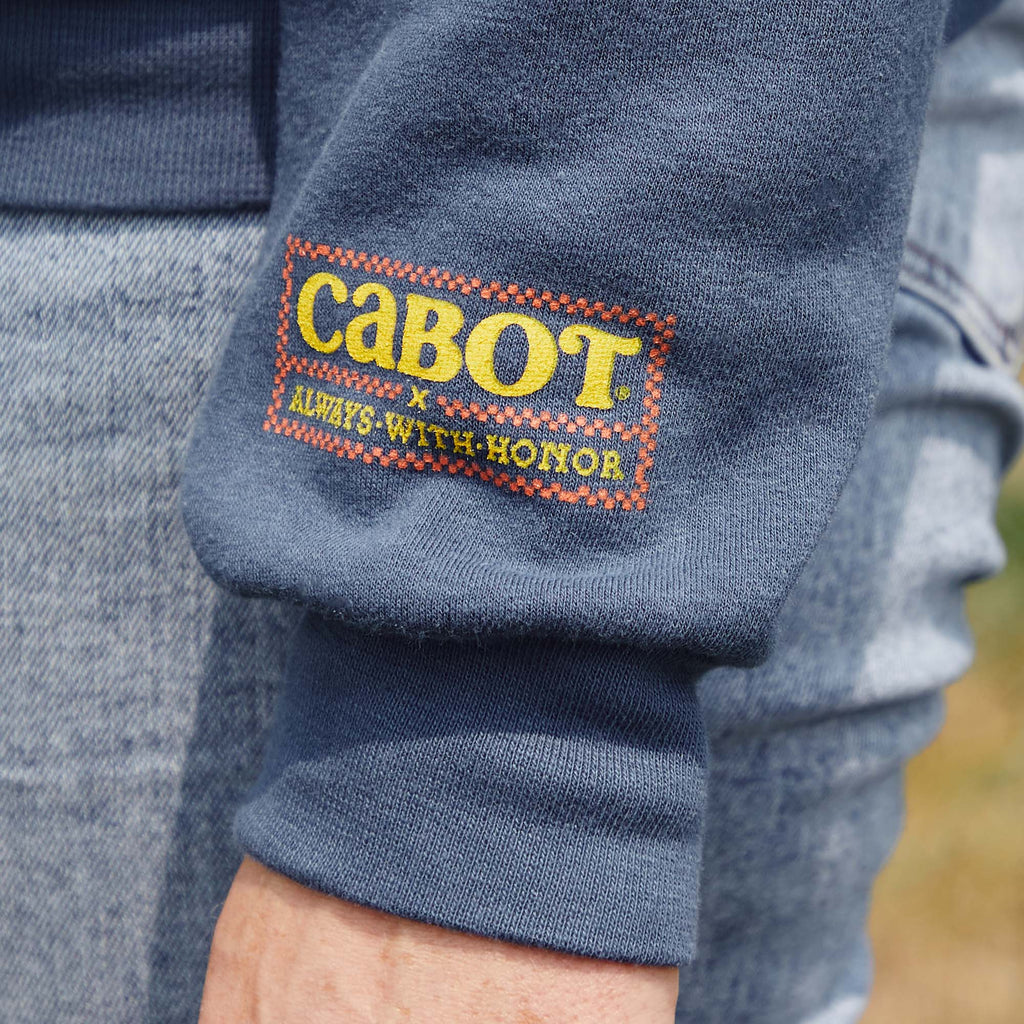 A close-up of the New Duds Cabot x Always With Honor Crewneck shows a blue sleeve with a stitched CABOT x ALWAYS WITH HONOR patch in yellow, red, and green. A hand and part of light blue jeans are also visible.