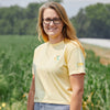 A woman with long blonde hair and glasses smiles outdoors in front of a green field, wearing the New Duds Cabot x Always With Honor Crop T-shirt. Trees and a road can be seen in the background.