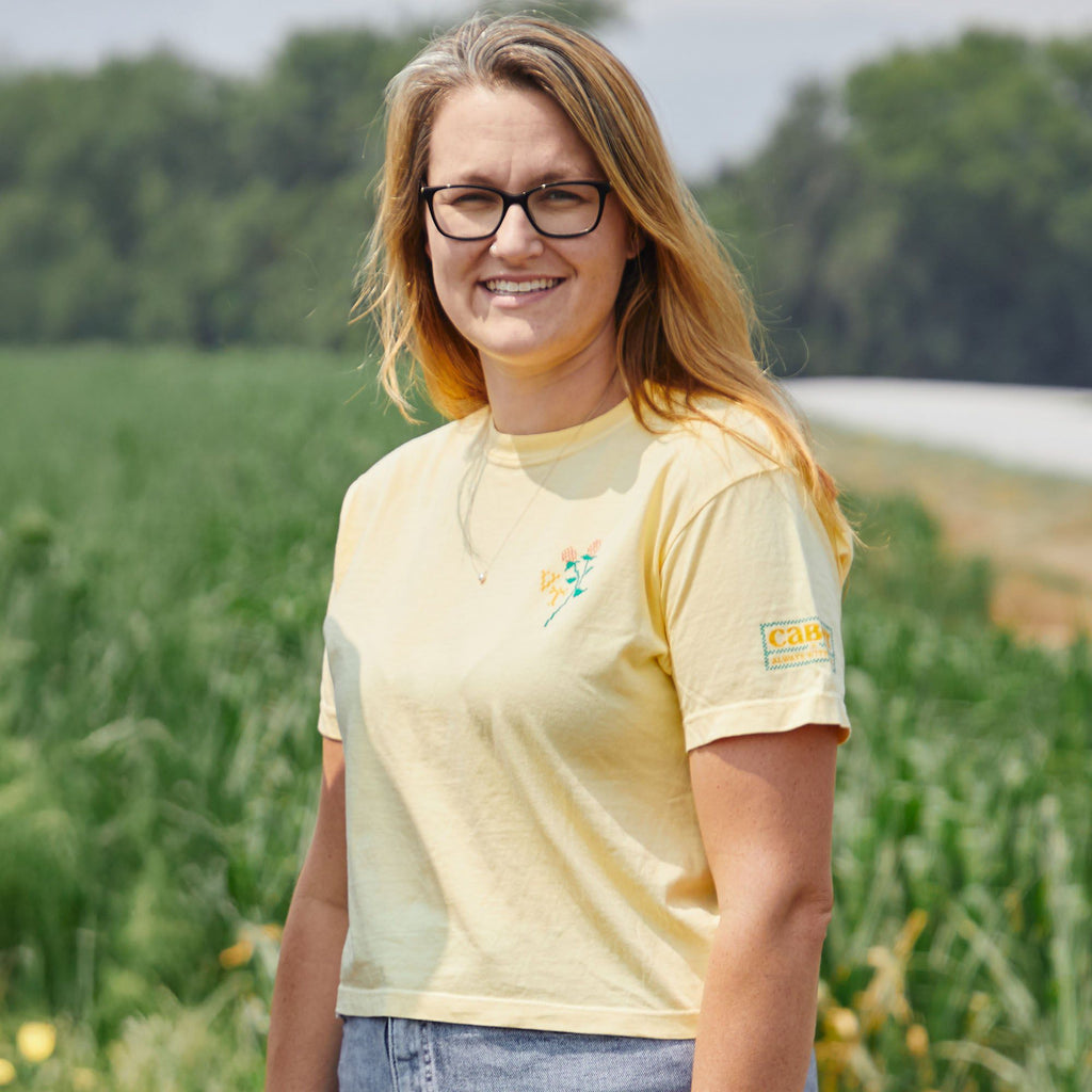 A woman with long blonde hair and glasses smiles outdoors in front of a green field, wearing the New Duds Cabot x Always With Honor Crop T-shirt. Trees and a road can be seen in the background.