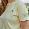 A person with long reddish-brown hair and a silver necklace is wearing the New Duds Cabot x Always With Honor Crop T-shirt, featuring a pale yellow color, stitched flower and VT on the chest, and a CABIN patch on the sleeve.