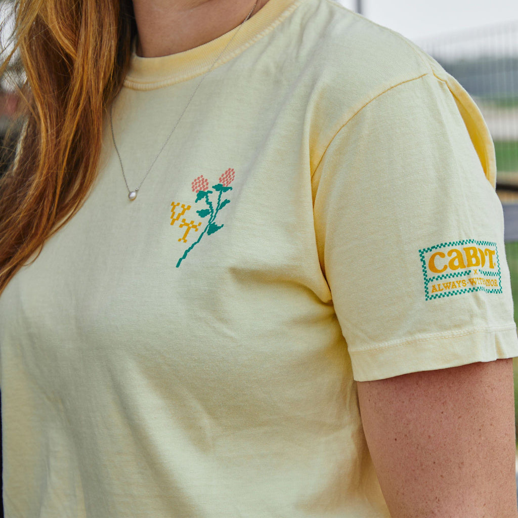 A person with long reddish-brown hair and a silver necklace is wearing the New Duds Cabot x Always With Honor Crop T-shirt, featuring a pale yellow color, stitched flower and VT on the chest, and a CABIN patch on the sleeve.