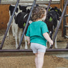 A young child with curly hair, wearing a Cabot x Always With Honor Toddler T-shirt by New Duds and gray shorts, stands at a farm pen watching black and white cows with yellow ear tags behind metal bars.