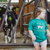 A young child wearing a green Cabot x Always With Honor Toddler T-shirt by New Duds stands in front of a metal-gated pen with black and white cows sporting yellow ear tags, gazing off to the side.