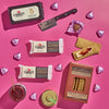 The Cabot Creamery Salty & Sweet Gift Pack features cheese bars, a small knife, crackers, jam, honey, and graham crackers on a pink backdrop with foil-wrapped heart chocolates—perfect for a delicious cheese and chocolate pairing.