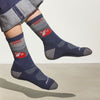 A person in rolled-up jeans and Darn Tough Vermont Cabot x Darn Tough Socks, Eclipse—navy blue Merino wool socks with red and white cassette tape designs—appears mid-air against a plain background.