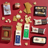 The Vermont Maple & Cheese Gift Box by Cabot Creamery features a flat lay of cheese, crackers, caramel, maple syrup with a winter scene tin, and Vermont maple candy on a red background.