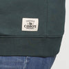 A close-up of the New Duds Vermont Sharp Chainstitched Crewneck, a relaxed-fit dark green sweatshirt with chain-stitched lettering and a white Cabot Creamery patch above the waistband, styled over blue jeans.