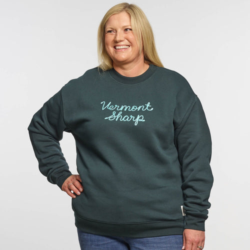 A smiling woman with long blonde hair poses against a plain background, wearing the New Duds Vermont Sharp Chainstitched Crewneck—a dark green relaxed-fit sweatshirt made from reclaimed plastic bottles, featuring chain-stitched lettering on the front.