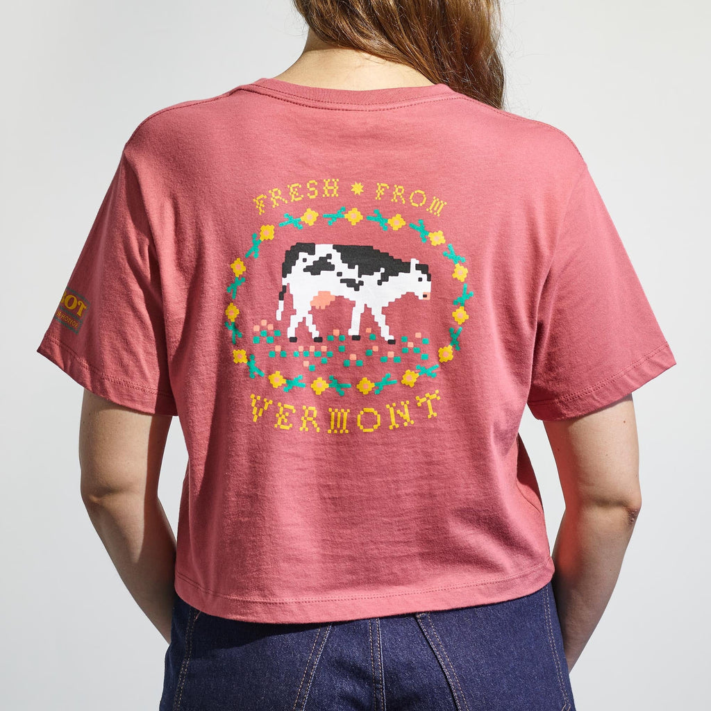 A person wearing the Cabot x Always With Honor Cropped T-shirt by New Duds—a pink, boxy fit shirt with a pixelated cow graphic, "Fresh From Vermont" on the back, and yellow text on the left sleeve.