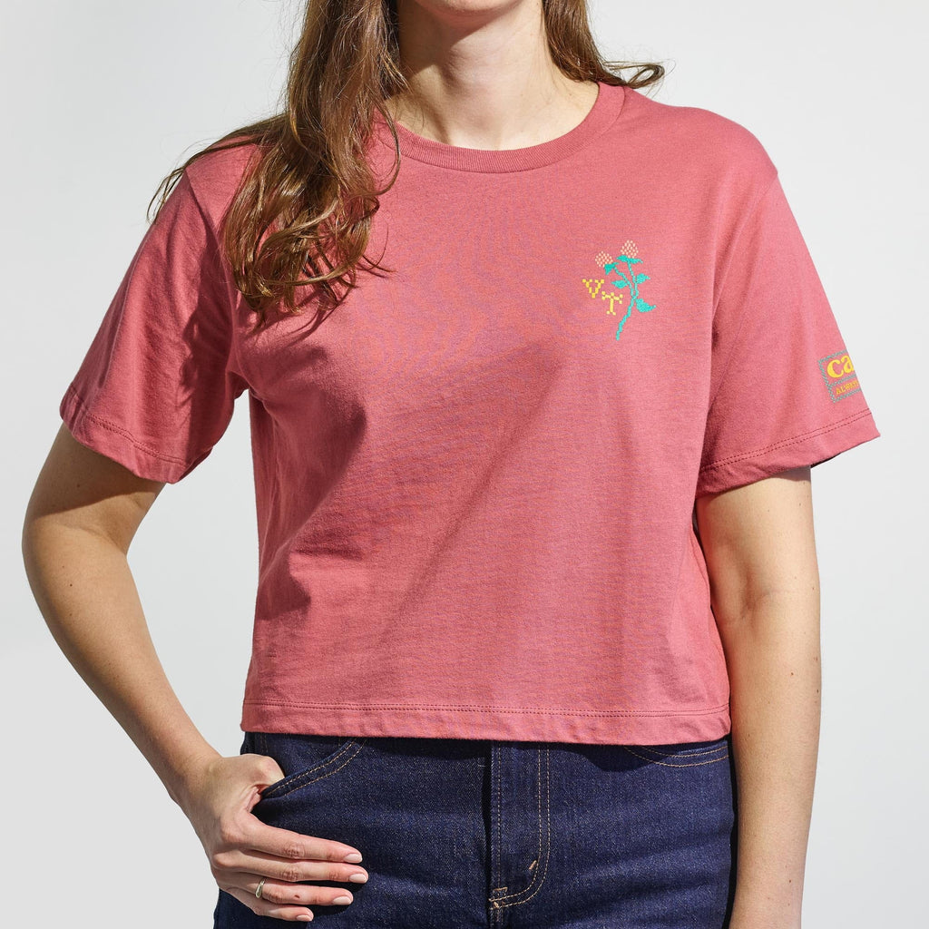 A woman wears the New Duds Cabot x Always With Honor Cropped T-shirt in coral pink, featuring a boxy fit and small abstract design on the left chest, paired with blue jeans as she stands with one hand in her pocket.