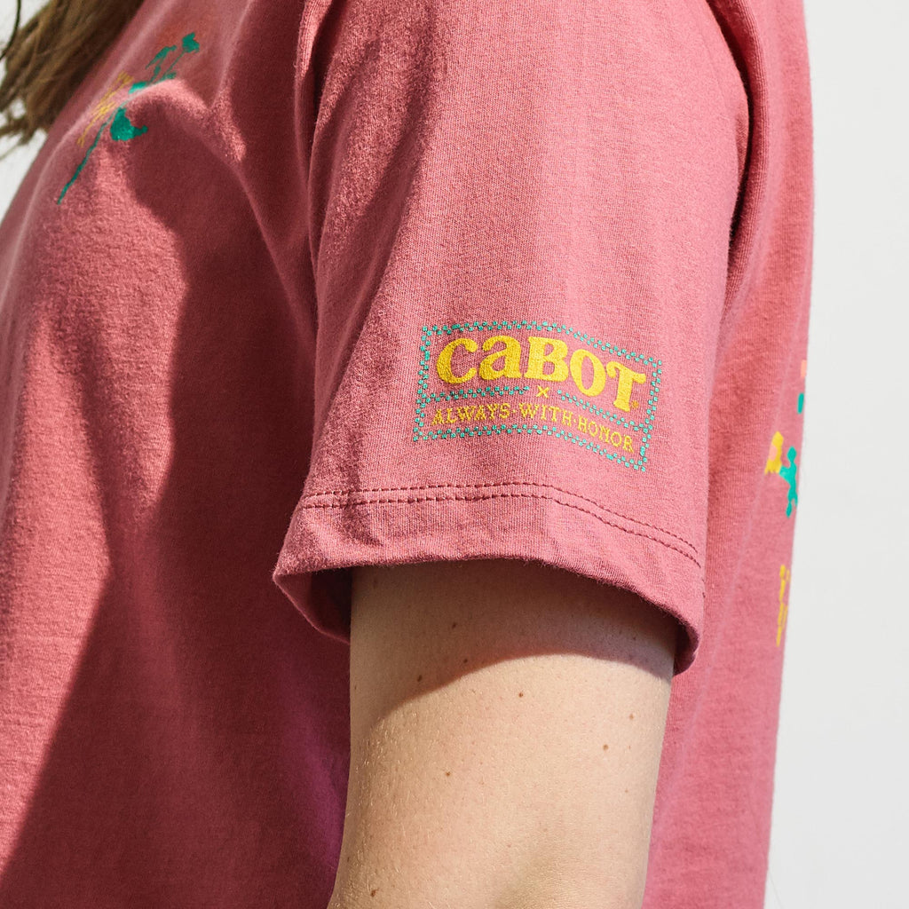 A person wears a rose Cabot x Always With Honor Cropped T-shirt by New Duds, made from ring-spun cotton. The boxy fit tee has a yellow and turquoise logo on the sleeve, with the image focused on the upper arm and side.
