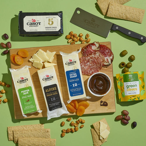 The Cabot Creamery X That Cheese Plate: Ultimate Charcuterie Gift Box features award-winning cheeses, dried apricots, salami slices, crackers, fig spread, and olives on a board with a cheese knife and extra crackers.
