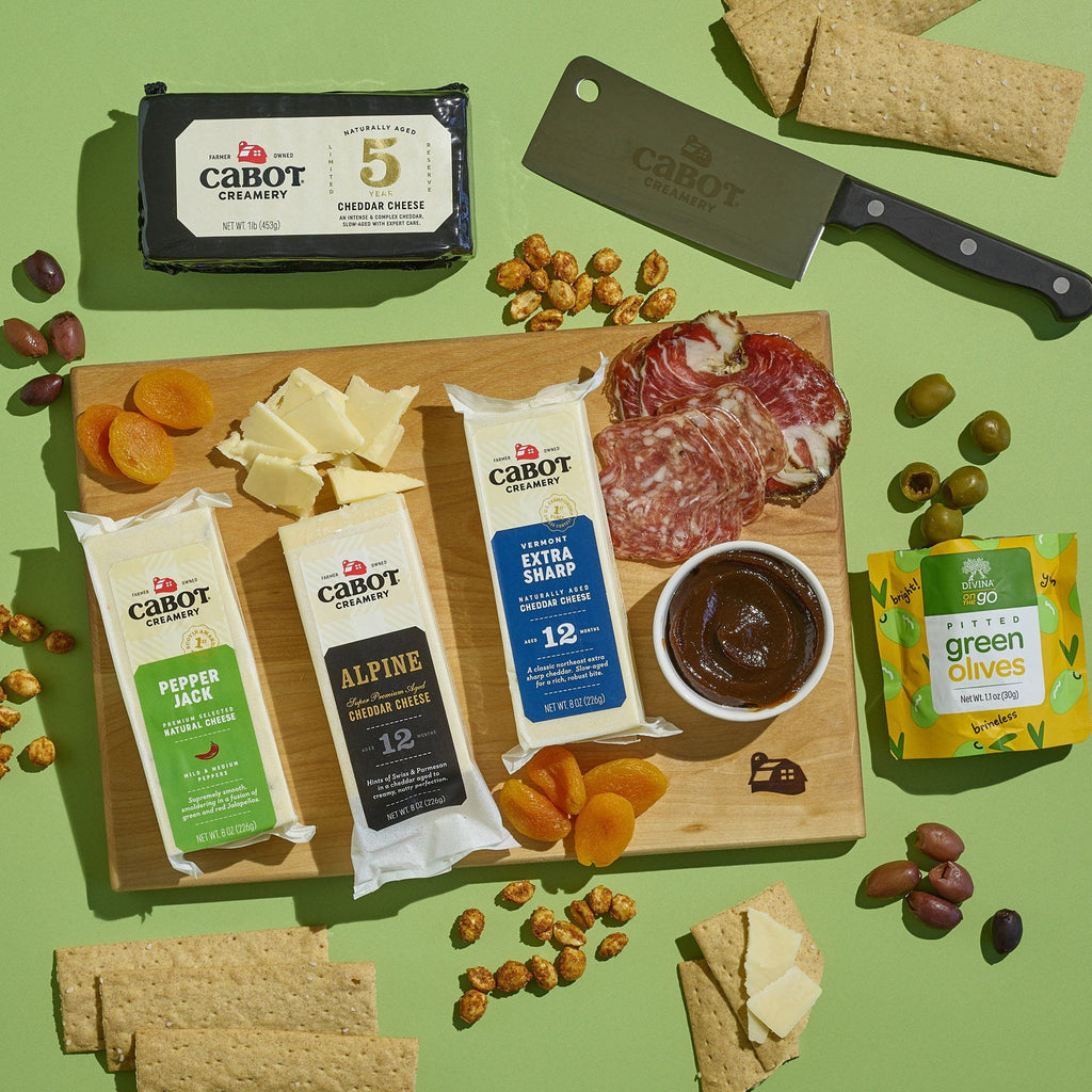 The Cabot Creamery X That Cheese Plate: Ultimate Charcuterie Gift Box features award-winning cheeses, dried apricots, salami slices, crackers, fig spread, and olives on a board with a cheese knife and extra crackers.