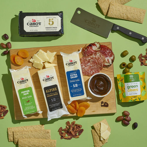 The Cabot X That Cheese Plate: Ultimate Charcuterie Gift Box by Cabot Creamery features cheeses, salami, dried apricots, dark jam, crackers, green olives, and a cheese knife on a wooden board against a green background.