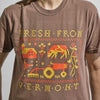 Person wearing the Cabot x Always With Honor T-shirt in Espresso by New Duds, featuring pixel art of a crab, tractor, plants, and "Fresh From Vermont" text in yellow and orange.