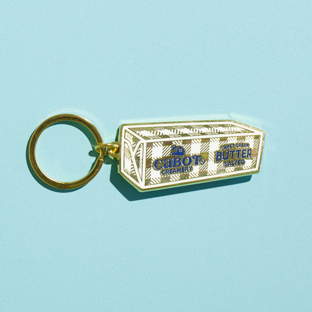 The WizardPins Butter Charm Keychain, shaped like a butter stick and crafted from hard enamel for a glossy finish, features a gold key ring and is displayed on a light blue background.