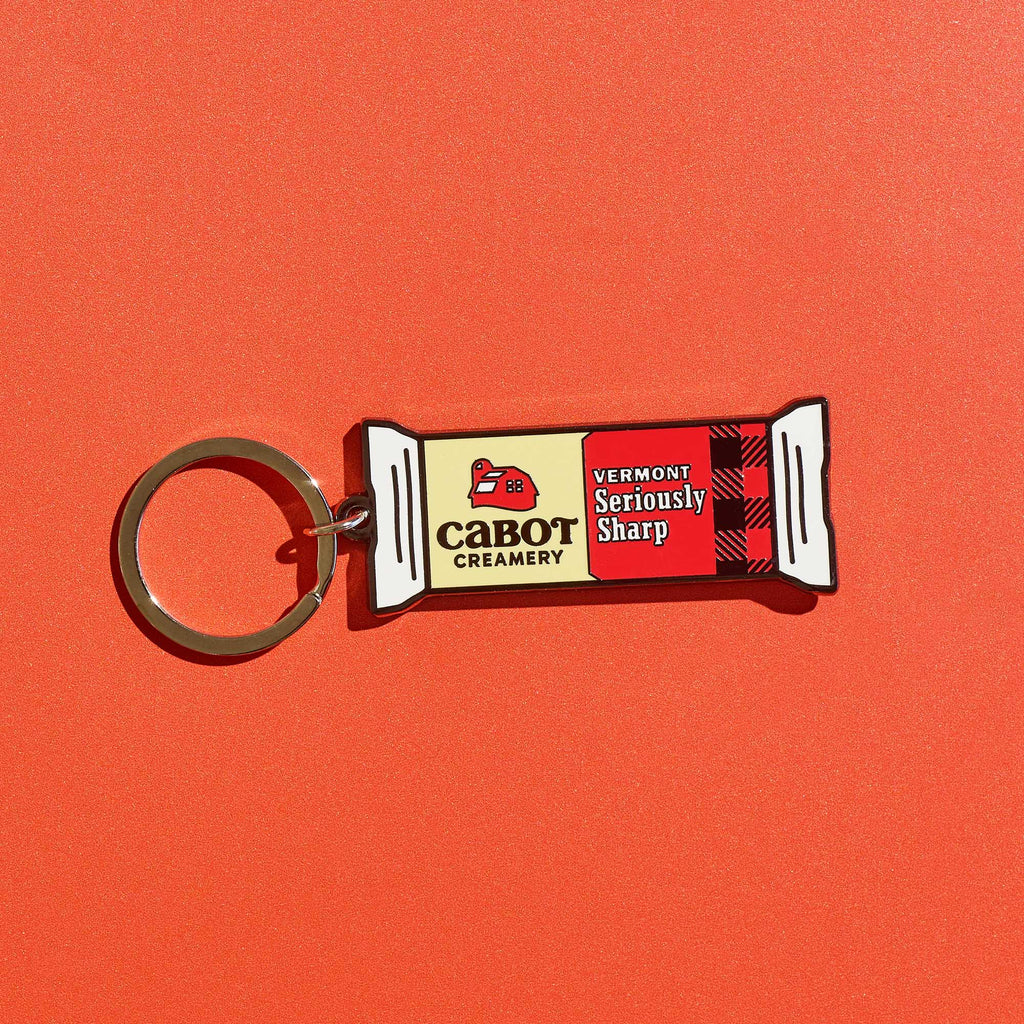 The WizardPins Seriously Sharp Charm Keychain, shaped like a cheese bar with “Cabot Creamery Vermont Seriously Sharp” label, stands out on a bold orange background.