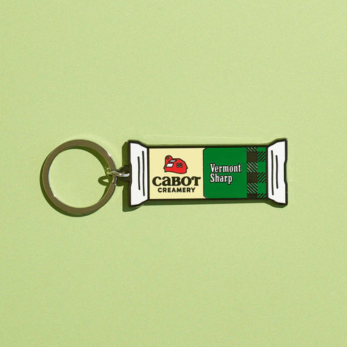 The Vermont Sharp Charm Keychain by WizardPins, inspired by Cabot Creamery's sharp cheddar packaging and logo, is shown on a light green background with its distinctive green and white design.