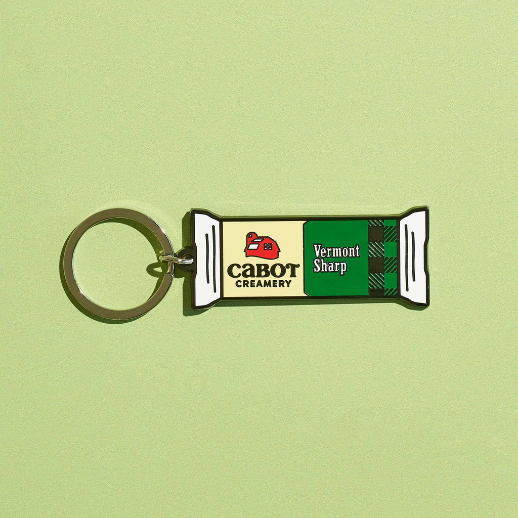 The Vermont Sharp Charm Keychain by WizardPins, inspired by Cabot Creamery's sharp cheddar packaging and logo, is shown on a light green background with its distinctive green and white design.