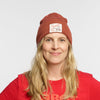 A smiling person with long blonde hair wears the New Duds “Life is Cheddar in Vermont Patch Beanie”—a red recycled polyester cuff beanie featuring a front white patch with text and an illustration—paired with a matching shirt.