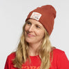 A smiling woman with long blonde hair wears the New Duds Life is Cheddar in Vermont Patch Beanie, a red recycled polyester cuff beanie with a white patch, and a red T-shirt against a plain light background.