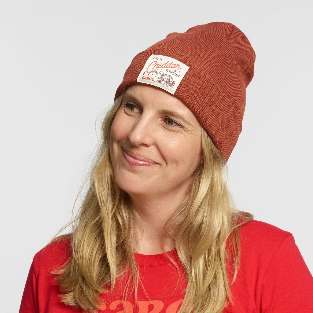 A smiling woman with long blonde hair wears the New Duds Life is Cheddar in Vermont Patch Beanie, a red recycled polyester cuff beanie with a white patch, and a red T-shirt against a plain light background.