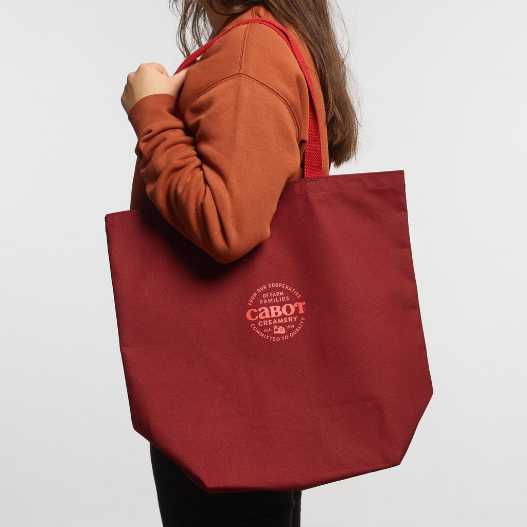 Wearing a rust sweatshirt, a person carries the New Duds Making the Cheddar Canvas Tote—a large maroon bag with red straps and a white Cabot Creamery logo, showing co-op pride—against a plain white background.