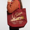 A person in a brown sweatshirt carries the New Duds Making the Cheddar Canvas Tote, which features a yellow cheese factory graphic and A Farmer Owned Cooperative text—perfect for showing true co-op pride.