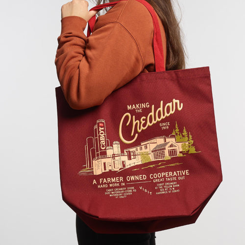A person in a brown sweatshirt carries the New Duds Making the Cheddar Canvas Tote, which features a yellow cheese factory graphic and A Farmer Owned Cooperative text—perfect for showing true co-op pride.