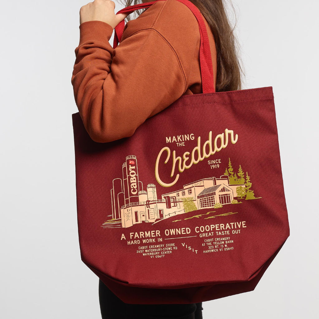 A person in a brown sweatshirt carries the New Duds Making the Cheddar Canvas Tote, which features a yellow cheese factory graphic and A Farmer Owned Cooperative text—perfect for showing true co-op pride.