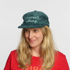 A woman with long blonde hair smiles against a white background, wearing a teal Vermont Sharp Chainstitched Corduroy Hat by New Duds and a red shirt.